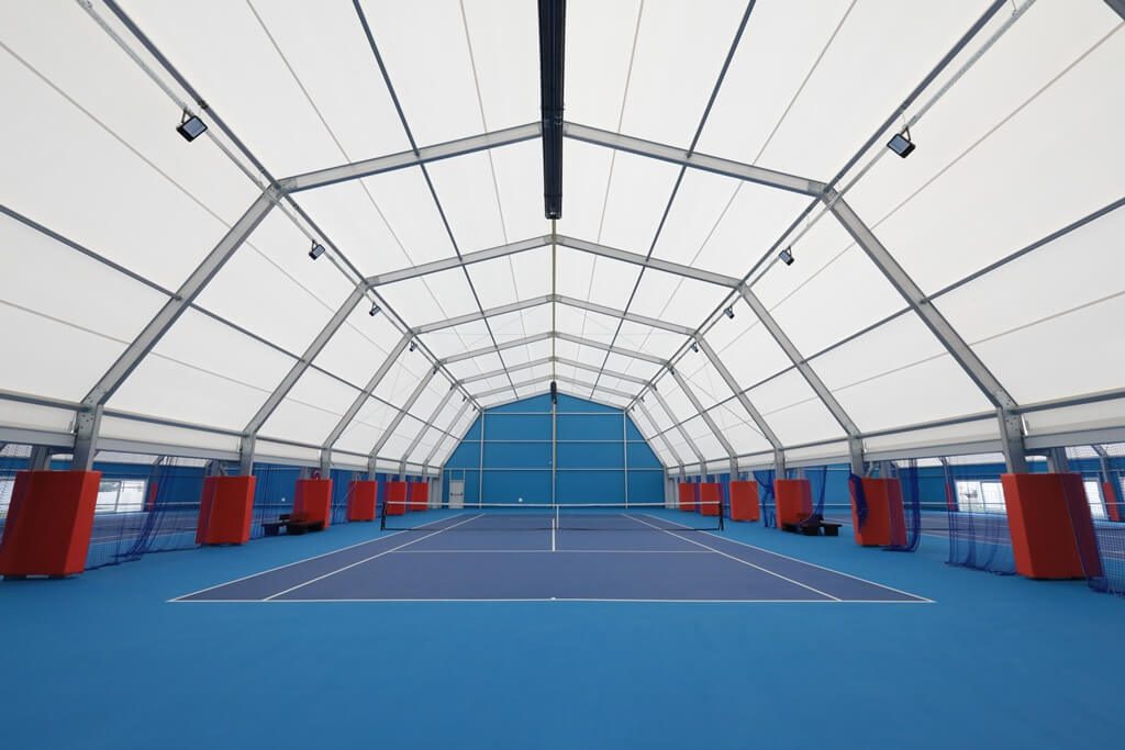 Interior view of a large clear span sports tent used as an indoor tennis court covering, featuring a heavy-duty aluminum frame and professional blue flooring.