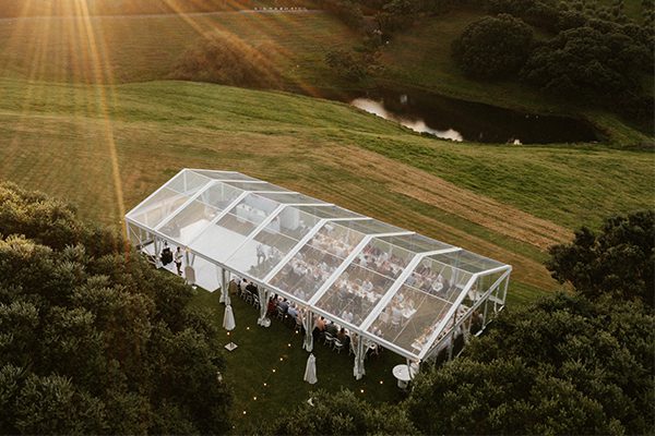 Aerial view of a luxury clear span wedding tent featuring a transparent PVC roof and white aluminum frame set up in a grassy field at sunset.