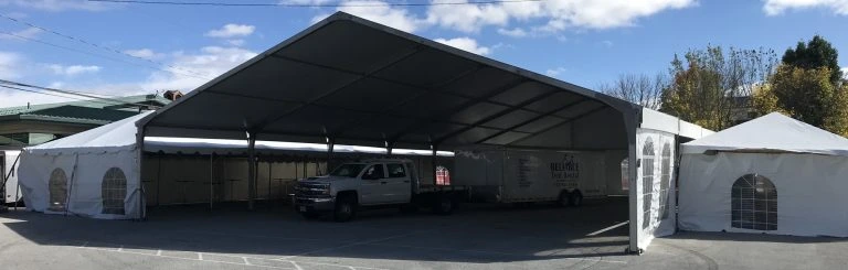 Large open-sided aluminum frame tent set up on an asphalt lot, sheltering a pickup truck and trailer parked underneath.