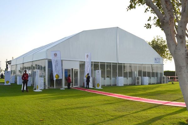 White polygon tent structure equipped with glass walls, double glass entrance doors, and external air conditioning units for a corporate event.